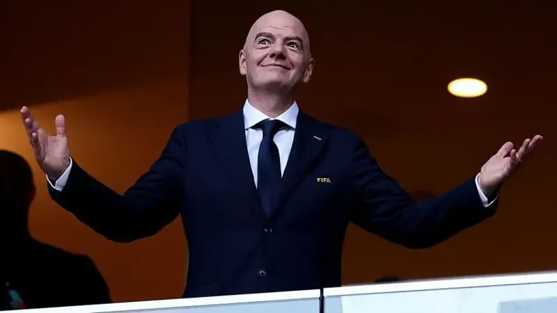 Gianni Infantino holds his arms out at each side with open palms, as he smiles while wearing a buttoned-up, dark smart jacket with FIFA written on its breast pocket